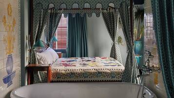 A bathroom with a bathtub, a bed with a floral quilt, and a window with curtains.