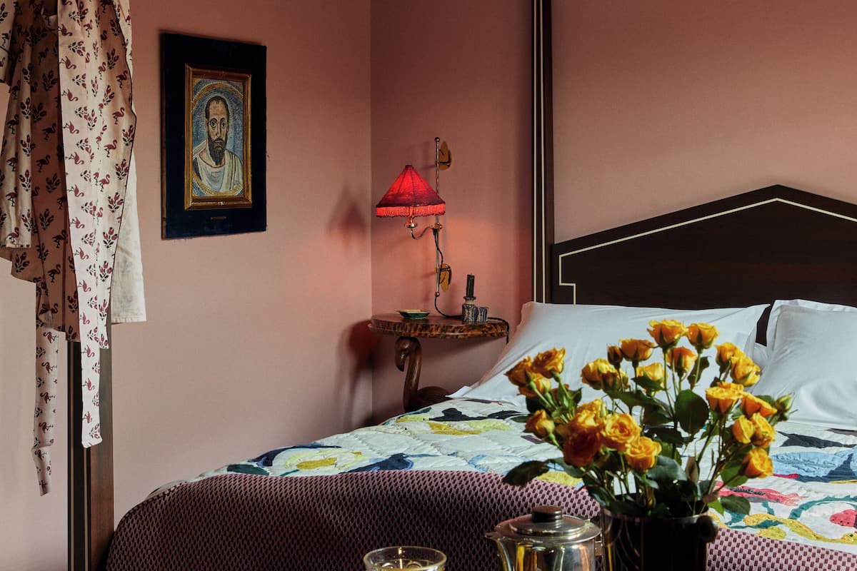 A hotel room with a bed, a table with breakfast items, a vase with flowers, and a lamp.
