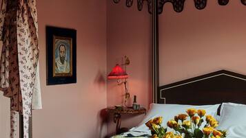 Una habitación de hotel con cama, una mesa con alimentos para el desayuno, un florero con flores y una lámpara.