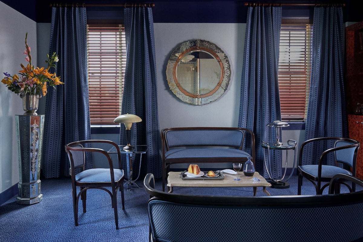 A room with a blue patterned carpet, a sofa, two chairs, a table with a vase of flowers, and a mirror on the wall.