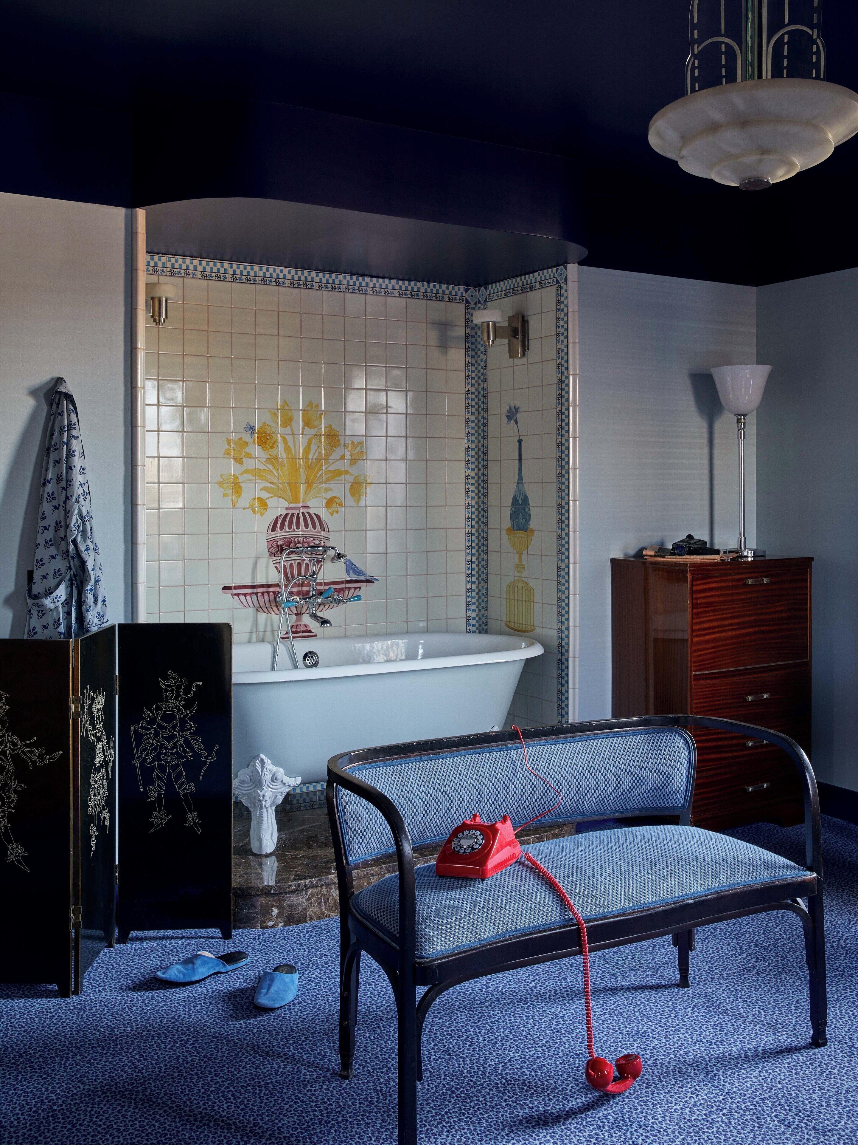 A bathroom with a bathtub, a sink, and a mirror. There is a blue patterned rug on the floor and a red telephone on the floor.