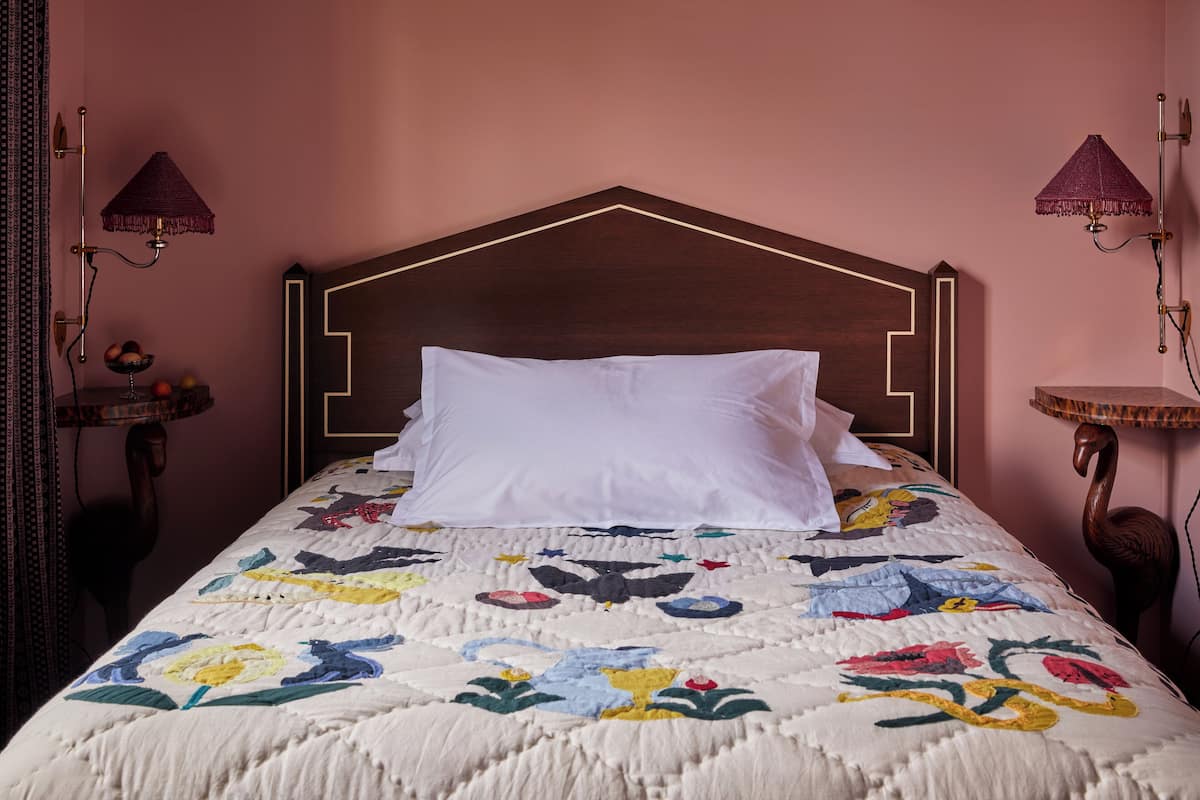 A bed with a floral quilt, a wooden headboard, and two wall-mounted lamps.