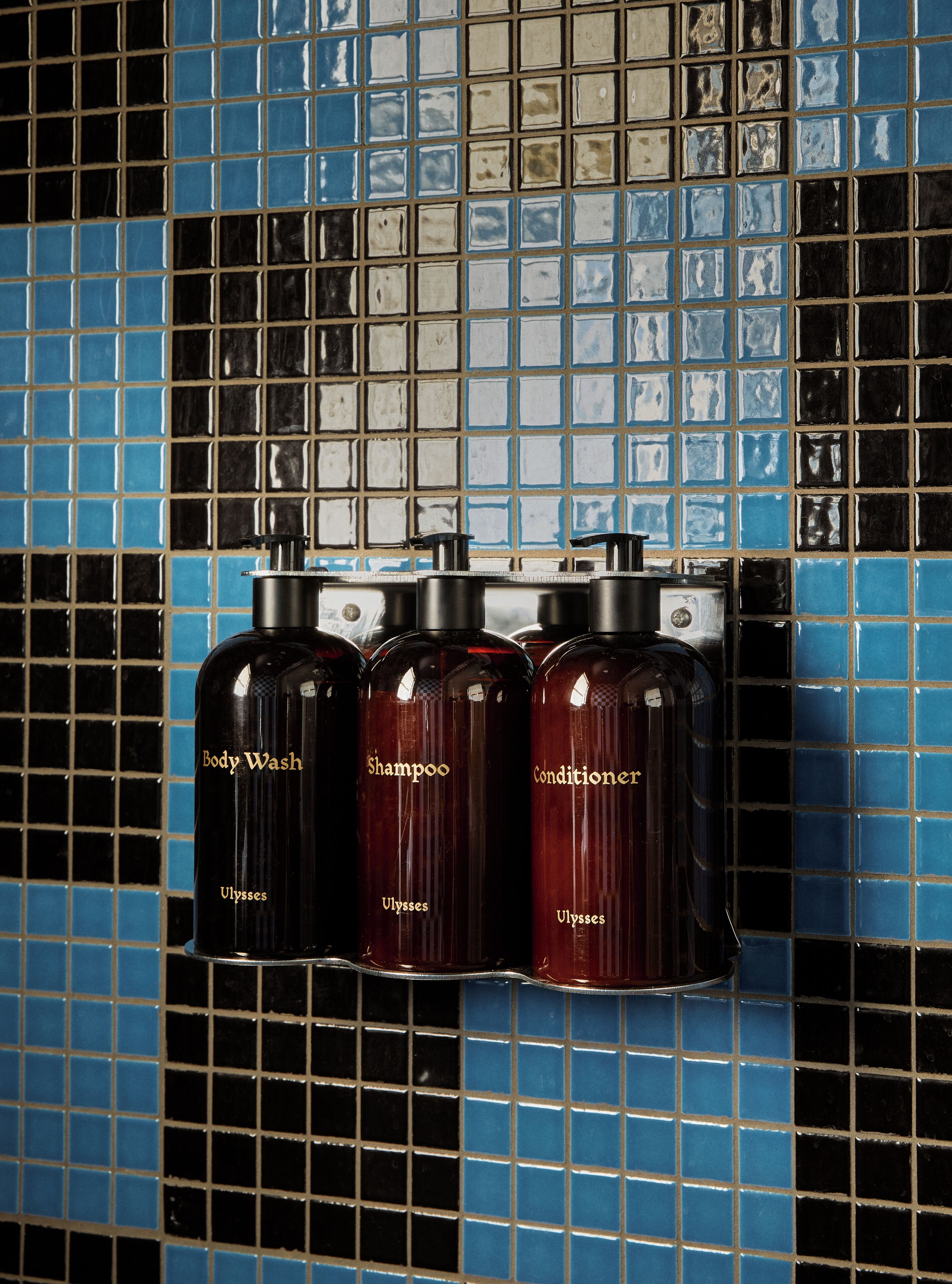Tres botellas de jabón líquido, champú y acondicionador Ulysses fijadas en una pared de azulejos.