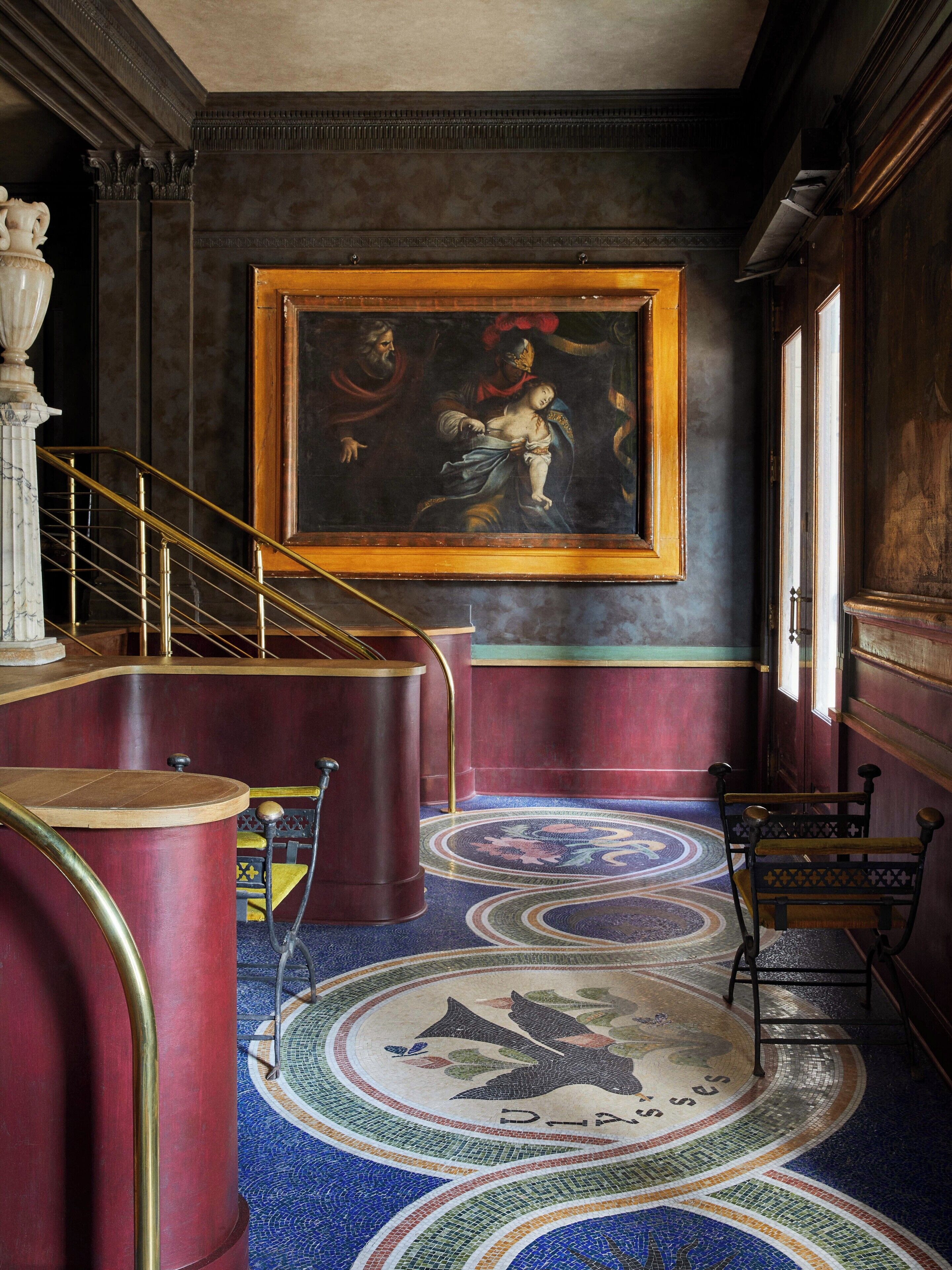 A grand hallway with a large classical painting, a marble staircase, and a detailed mosaic floor.