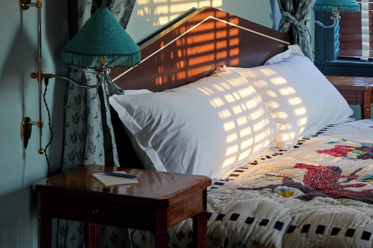 A bedroom with a wooden headboard, a bedside table with a lamp, and curtains allowing sunlight to filter through.