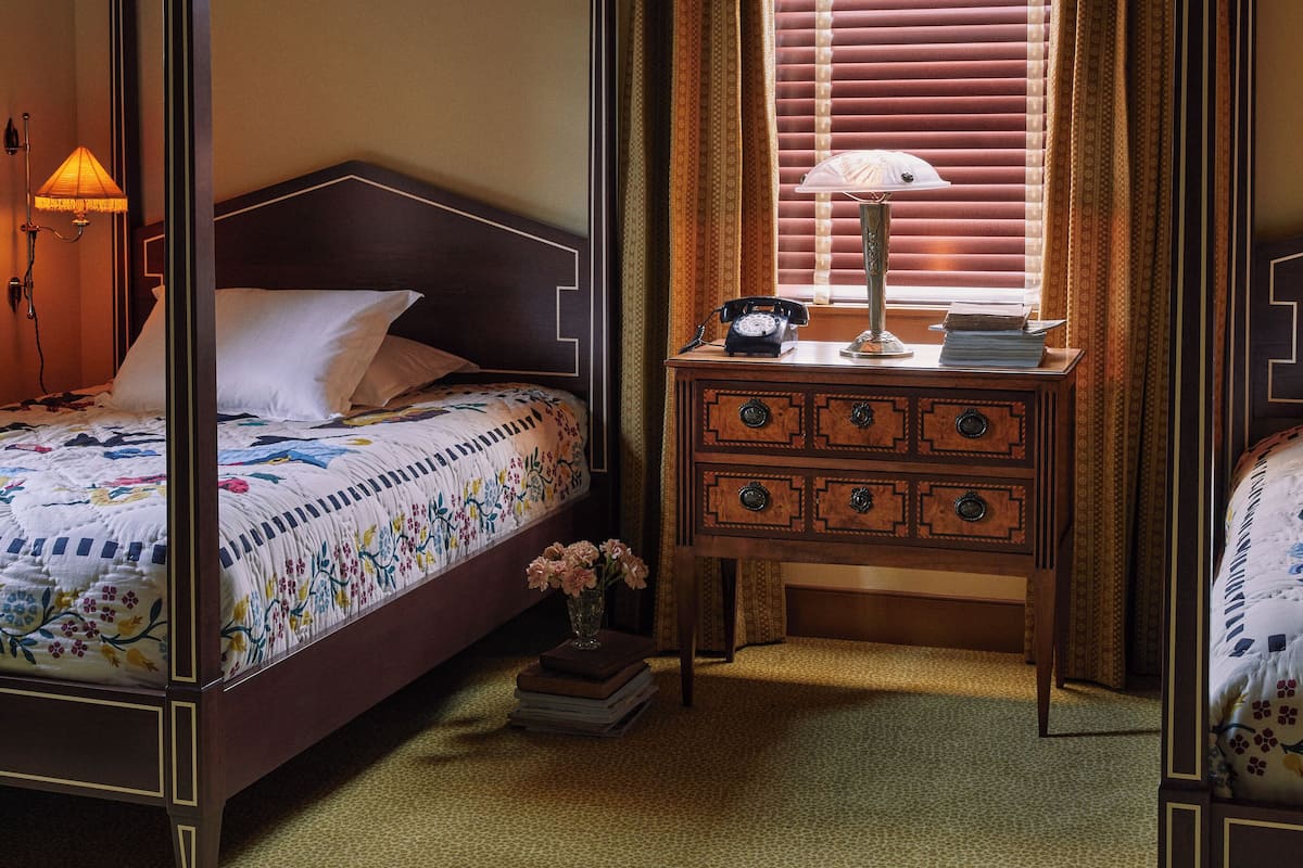 A four-poster bed with a floral bedspread, a bedside lamp, a wooden nightstand with drawers, and a window with blinds.