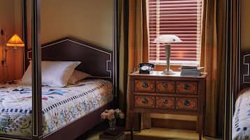 A four-poster bed with a floral bedspread, a bedside lamp, a wooden nightstand with drawers, and a window with blinds.