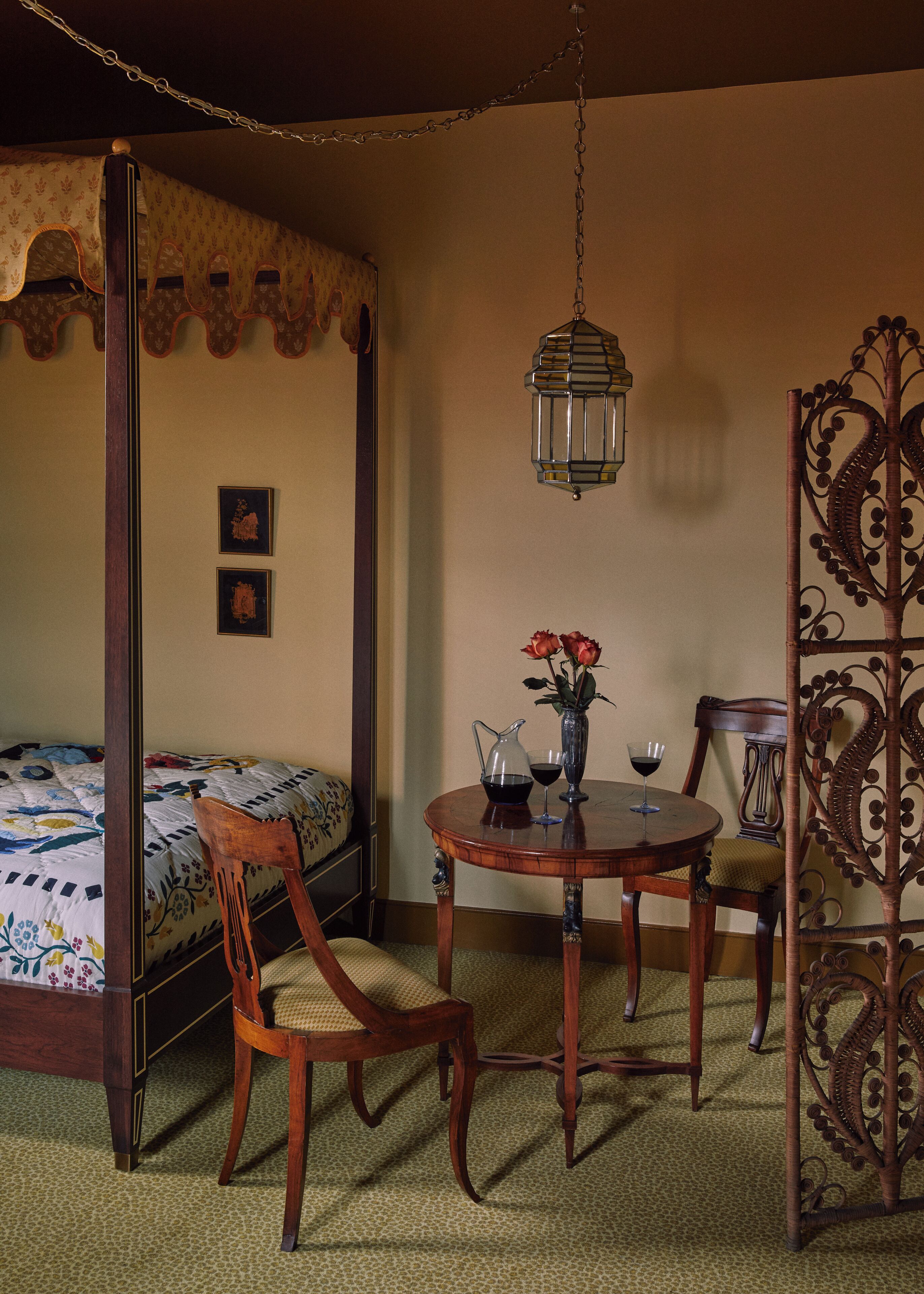 Een hemelbed met bloemenbedekking, een houten stoel met een geweven zitvlak, een ronde houten tafel met een bloevas en twee wijnglazen, en een hangende lantaarn.