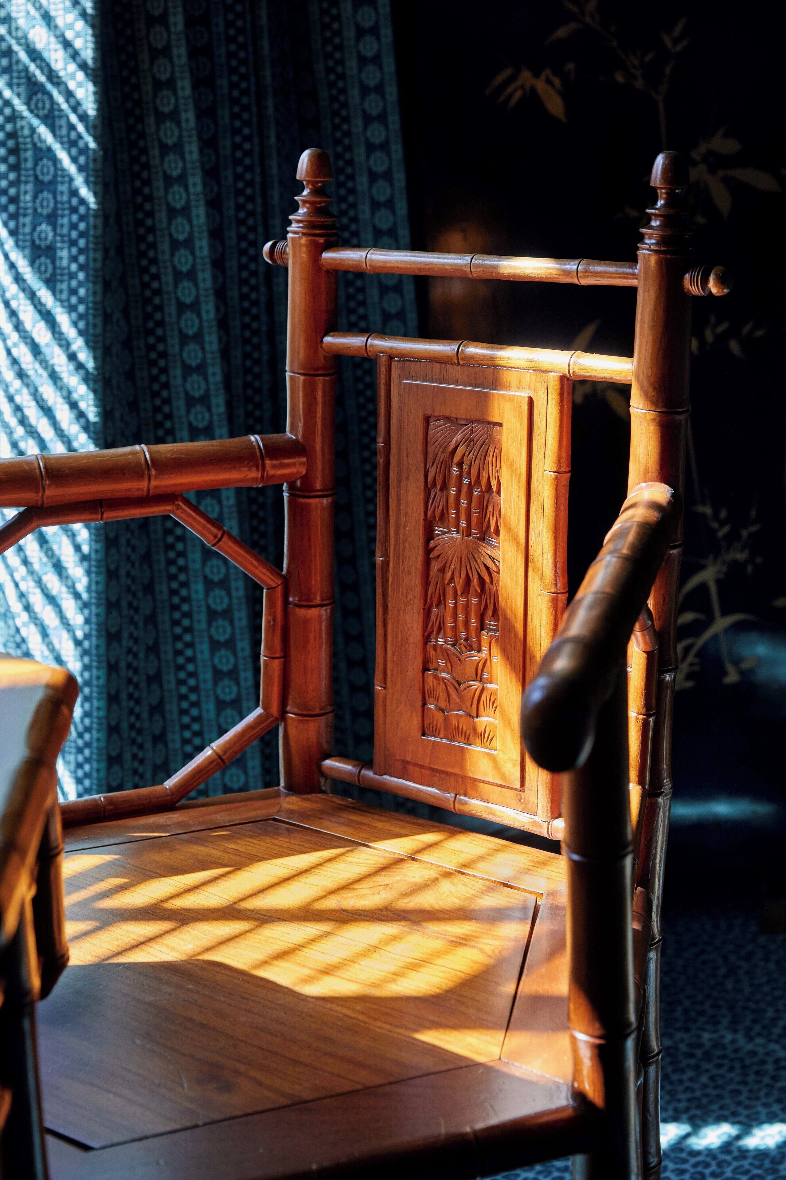 Une chaise en bois, dotée d’un dossier sculpté et d’une assise en osier, placée dans une pièce où l’on aperçoit des rideaux à motifs et une plante en arrière-plan.