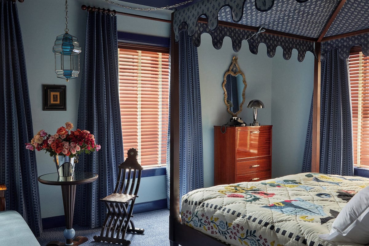 A bedroom with a four-poster bed, a dresser, a chair, a table with a vase of flowers, and a hanging lantern.