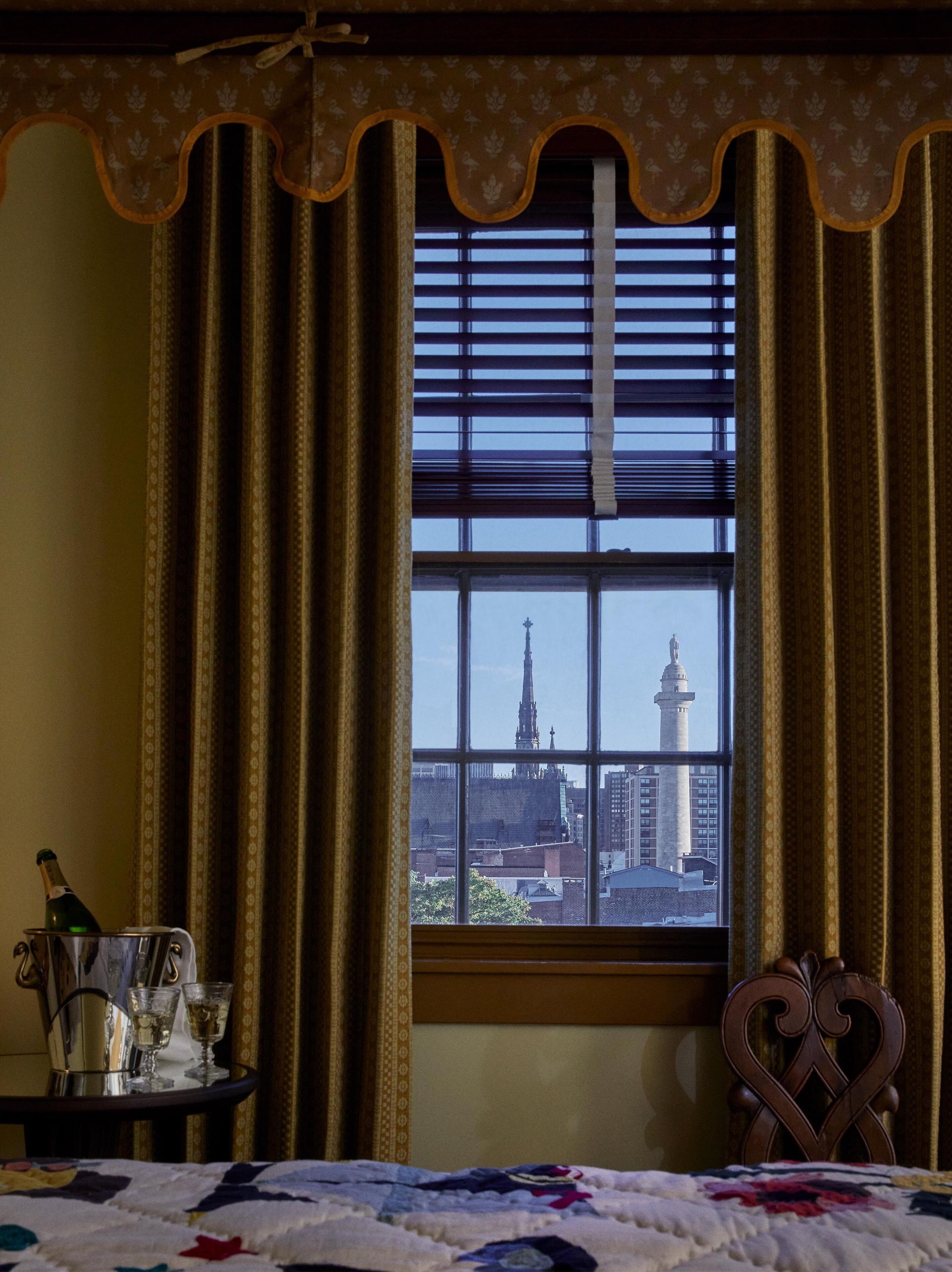 A room with a large window, curtains, a bed, a bedside table with a bottle and glasses, and a decorative chair.