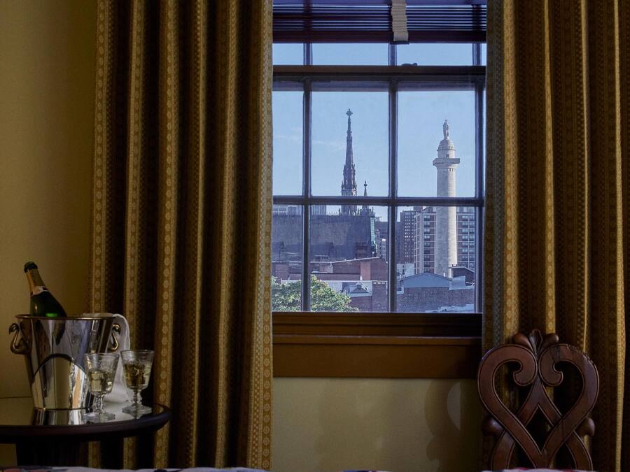 Ein Zimmer mit einem großen Fenster, Vorhängen, einem Bett, einem Nachttisch mit Flasche und Gläsern sowie einem dekorativen Sessel.