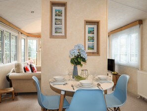 A dining area with a round table set for four, surrounded by blue chairs. There are framed pictures on the wall, a vase with flowers, and a television on a stand.