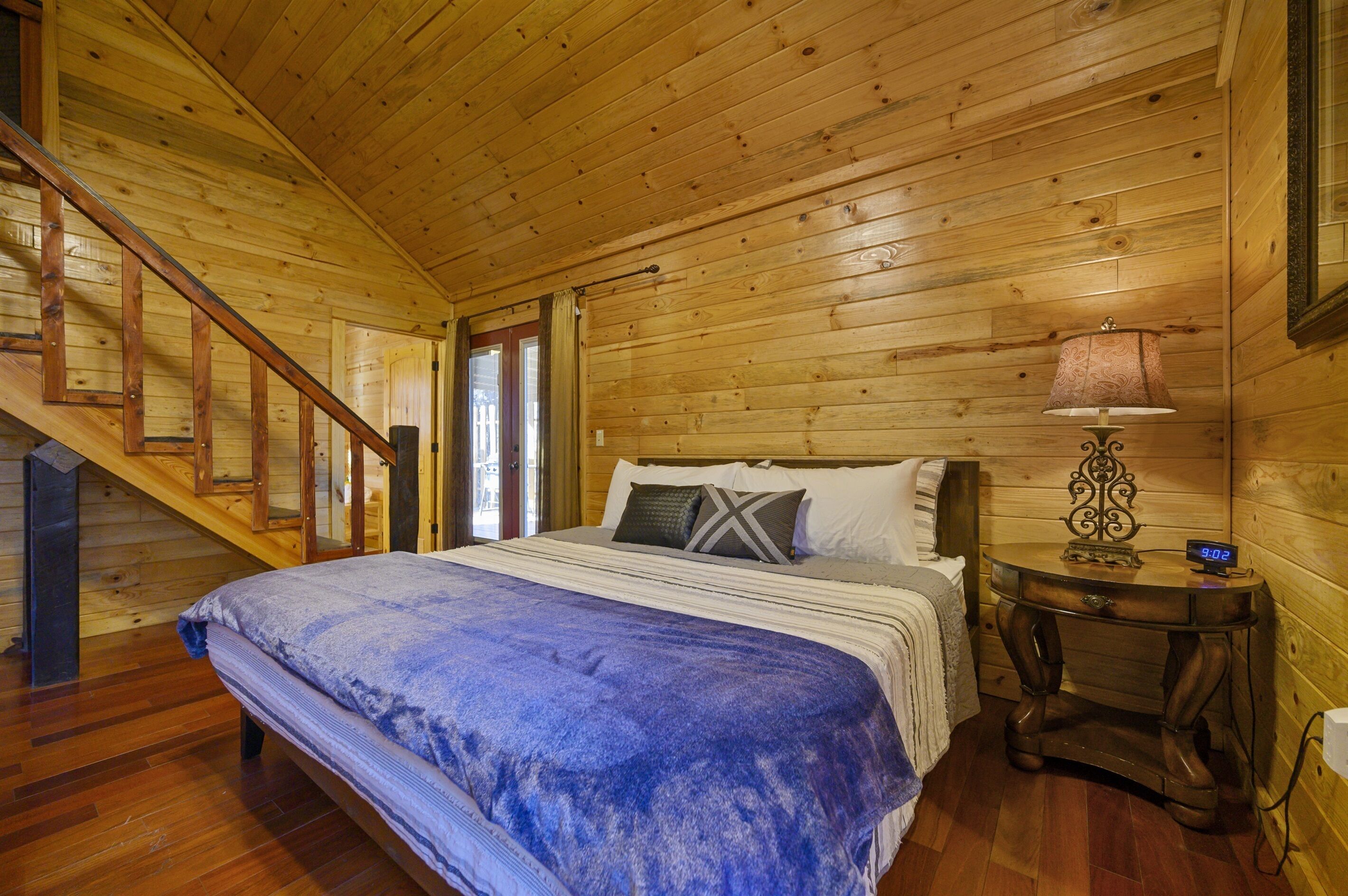 Een slaapkamer in een houten hut met een bed, een nachtkastje, een lamp en een trap.
