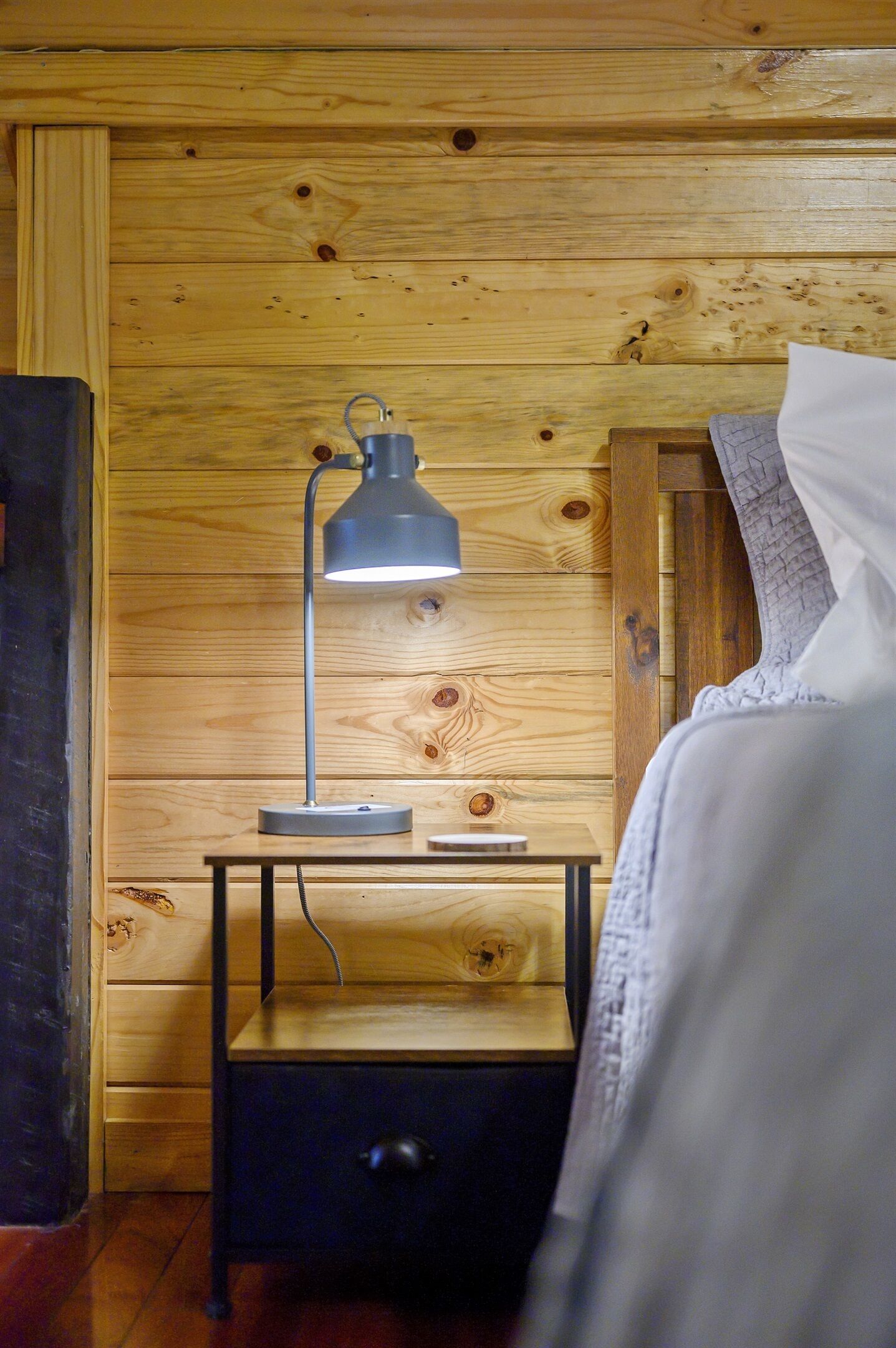 Une chambre de style cabane en bois, avec une seule table de chevet, une lampe et un lit recouvert de linge de lit blanc.