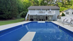 Piscina retangular com um deck branco, cercada por uma área gramada e uma casa com varanda.