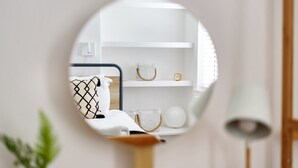 Um espelho redondo reflete um quarto com uma cama, mesa de cabeceira, abajur e prateleiras com objetos decorativos.