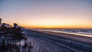 Une plage au coucher du soleil, avec des bâtiments en arrière-plan et des empreintes de pas dans le sable.