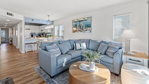 Un salon moderne avec un canapé d’angle gris, une table basse en bois et un tableau représentant une scène de plage.