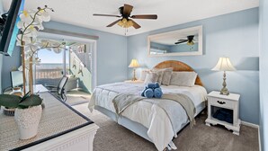 Une chambre à coucher avec un lit, deux tables de chevet avec des lampes, un ventilateur de plafond, un téléviseur fixé au mur et une grande fenêtre donnant sur un beau paysage.