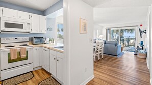 Une cuisine moderne avec des meubles blancs, un four à micro-ondes et une cuisinière. On y trouve un coin repas avec un canapé gris et une table. La pièce est dotée d’un plancher en bois et de grandes fenêtres.