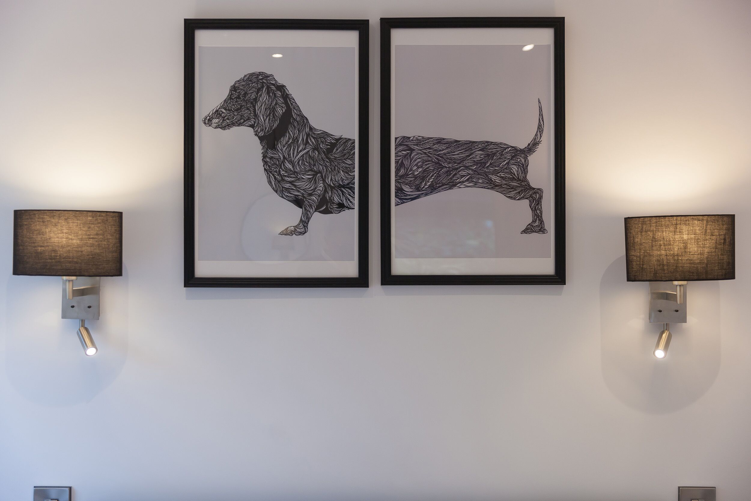 Two framed black and white dog illustrations on a wall, flanked by wall-mounted lamps.