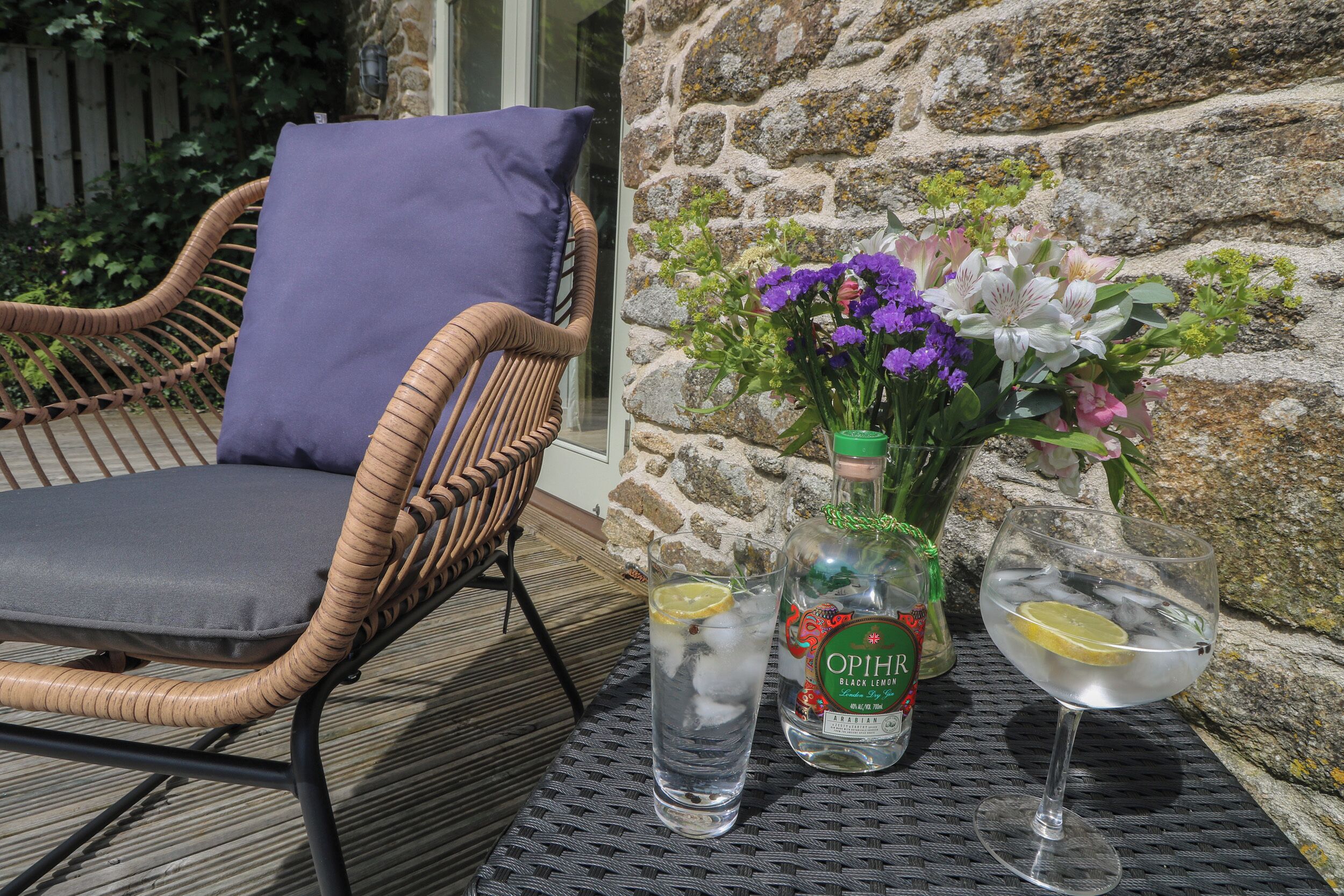 A wicker chair with a cushion, a bottle of OPIHER Black Lemon, a glass with a lemon slice, and a vase with flowers on a wooden deck.