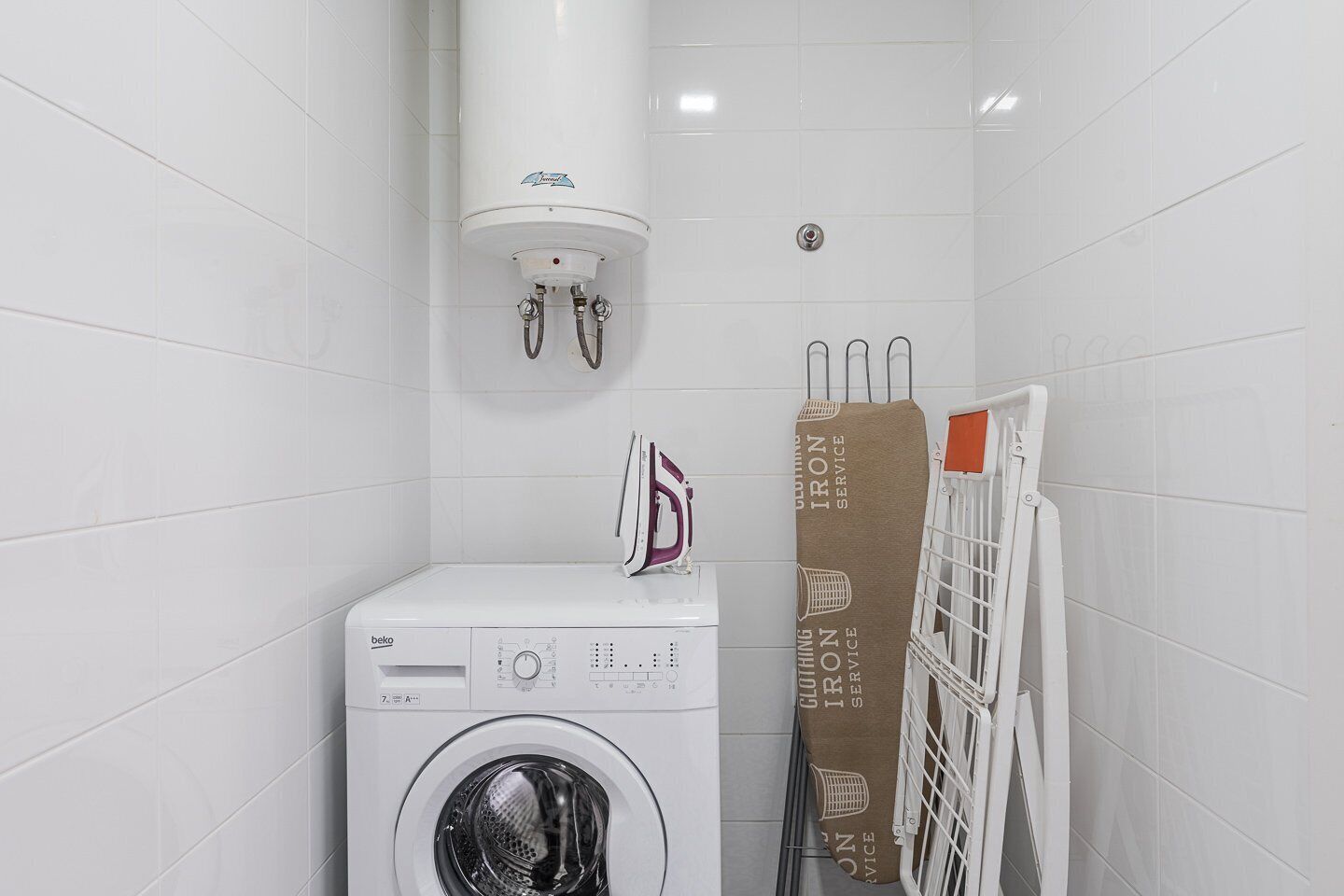 A compact laundry room with a front-loading washing machine, a wall-mounted heater, and a drying rack.