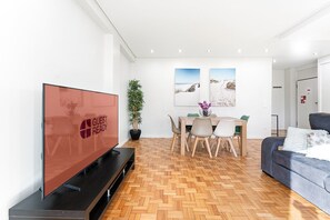Un salon moderne avec un téléviseur à écran plat, un coin repas avec une table et des chaises en bois, une plante en pot et un canapé bleu.
