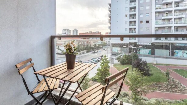 A balcony with a wooden table and chairs, a potted plant, and a view of a cityscape with buildings and a road. - GuestReady - Broken Record with Balcony + Parking (Porto)