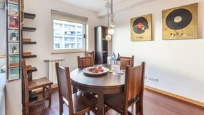 A dining area with a round table, chairs, a plate of food, a water bottle, and a bottle of wine. There are framed pictures on the wall and a window with a view of buildings. - GuestReady - Broken Record with Balcony + Parking (Porto)