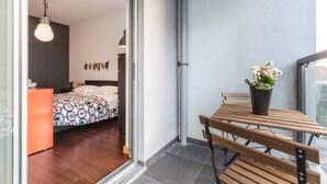 A balcony with a wooden table and chairs, a potted plant, and a view of a bedroom with a patterned bedspread and a hanging light fixture. - GuestReady - Broken Record with Balcony + Parking (Porto)