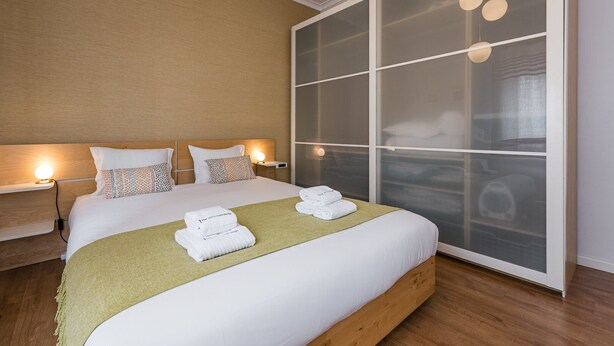 A modern hotel room with a large bed, two bedside tables with lamps, a large wardrobe with frosted glass doors, and a green throw blanket on the bed. - GuestReady Essential - Heartbeats (Porto)