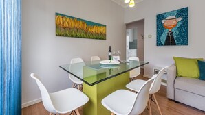 A modern dining area with a glass table, white chairs, and a green accent wall. There are two paintings on the walls, one of yellow flowers and another of a person in a hat. - GuestReady Essential - Heartbeats (Porto)