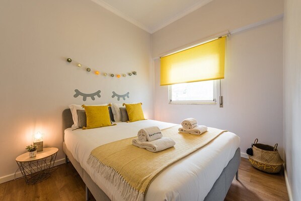 A neatly arranged hotel room with a bed, pillows, a bedside table, a lamp, a window with a yellow blind, and a small basket. - GuestReady Essential - Heartbeats (Porto)
