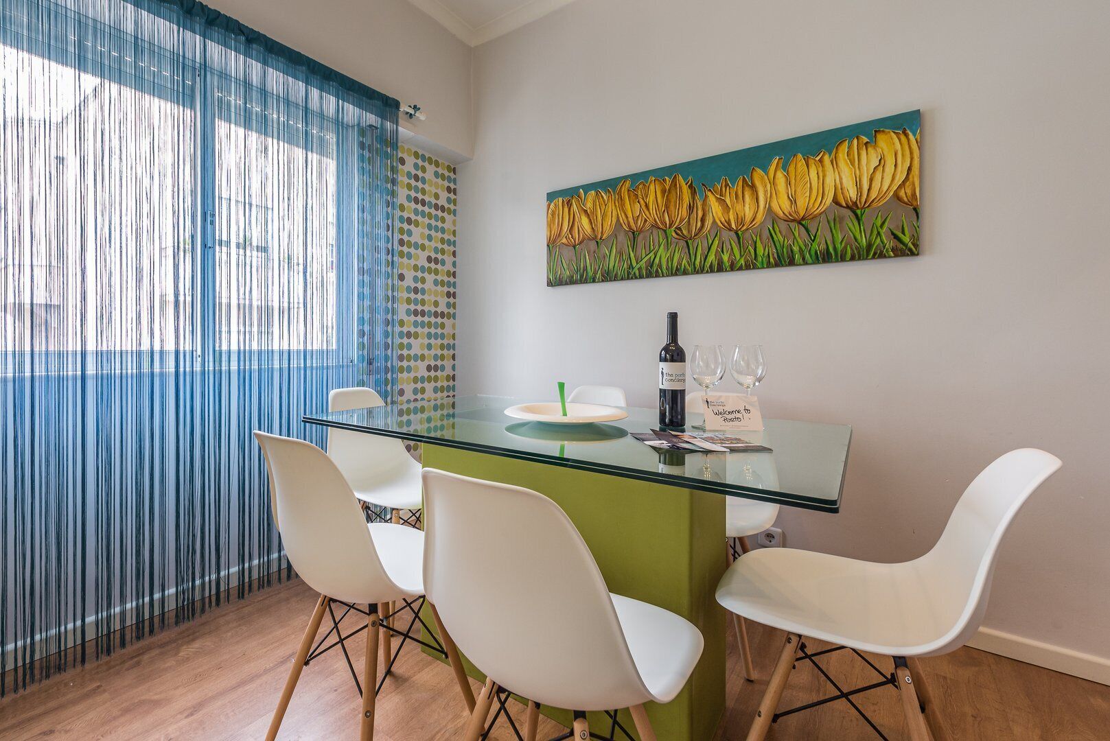 A dining area with a glass table, white chairs, a bottle of wine, and a painting of yellow flowers on the wall.