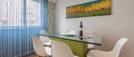 A dining area with a glass table, white chairs, a bottle of wine, and a painting of yellow flowers on the wall.