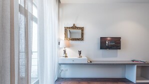 Ein Hotelzimmer mit Holzboden, einem weißen Schreibtisch mit Lampe, einem dekorativen Spiegel und einem an der Wand montierten Fernseher.