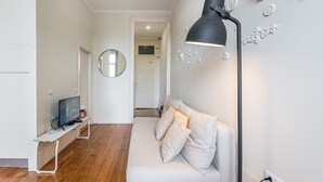 En moderne stue med en hvid sofa, en sort gulvlampe, et rundt spejl og et fjernsyn på et hvidt møbel.