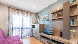 A modern living room with a flat-screen TV, wooden shelving, and a bed with a pink cover. - GuestReady - Sound of Nature - T1 Boavista (Porto)