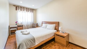 A neatly arranged hotel room with a wooden bed, a bedside table with a lamp, a nightstand, and a small desk with a chair. - GuestReady - Sound of Nature - T1 Boavista (Porto)