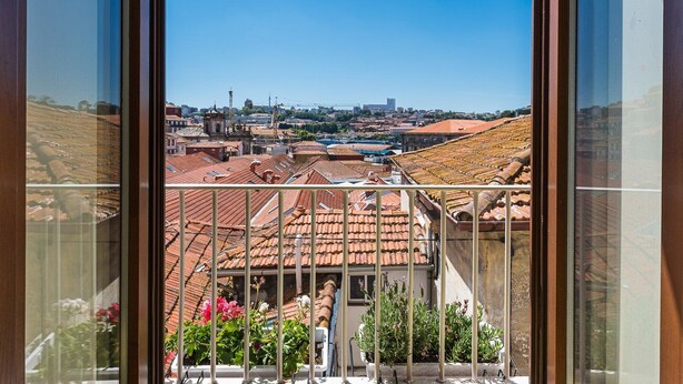 Blick von einem Balkon auf Terrakotta-Dächer, einem klaren blauen Himmel und eine ferne Stadtlandschaft.