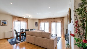 A spacious living area with a beige sofa, a dining table with chairs, and a large window with curtains. - GuestReady - Luxe living by the pool (Póvoa de Varzim)