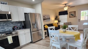 Una cocina moderna con electrodomésticos de acero inoxidable, un comedor con sillas blancas y un mantel amarillo, y una sala de estar con un sofá y un ventilador de techo.