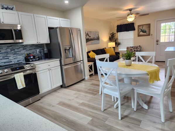 A modern kitchen with stainless steel appliances, a dining area with white chairs and a yellow tablecloth, and a living room with a sofa and a ceiling fan.