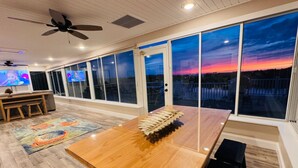 Ein geräumiges Zimmer mit einem großen Holztisch, einem Deckenventilator und Blick auf einen Sonnenuntergang durch große Fenster.