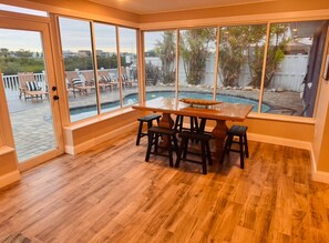 A room with a wooden floor, a table with four stools, and large windows overlooking a backyard with a pool and outdoor seating.