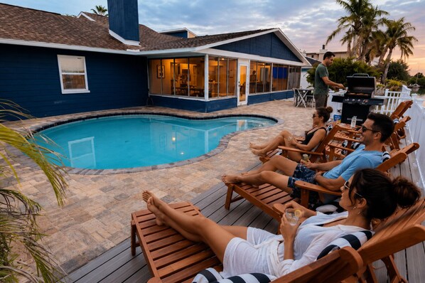 A poolside area with a pool, wooden deck, and a house with a large window overlooking the pool.