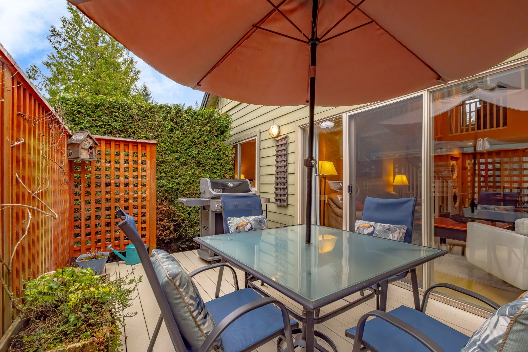 An outdoor patio with a glass table and chairs, a wooden fence, and a grill.