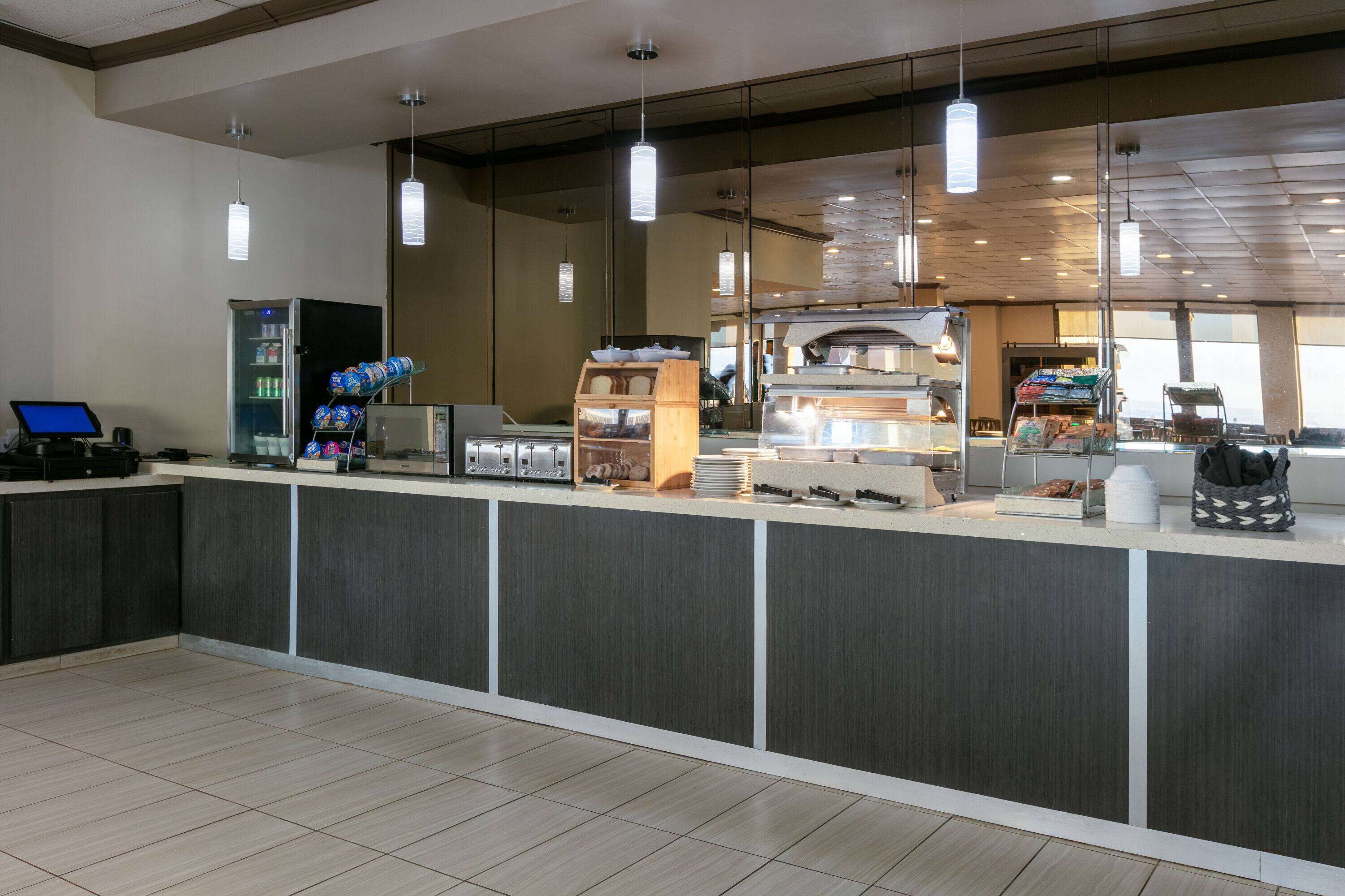 A modern hotel lobby with a reception desk, vending machines, a coffee station, and a large window offering a view of the city.